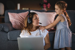 Front view of multitasking mother asking her active daughter not to interrupt her while she uses laptop computer and headset to work remotely. Little girl shows interest in playing.